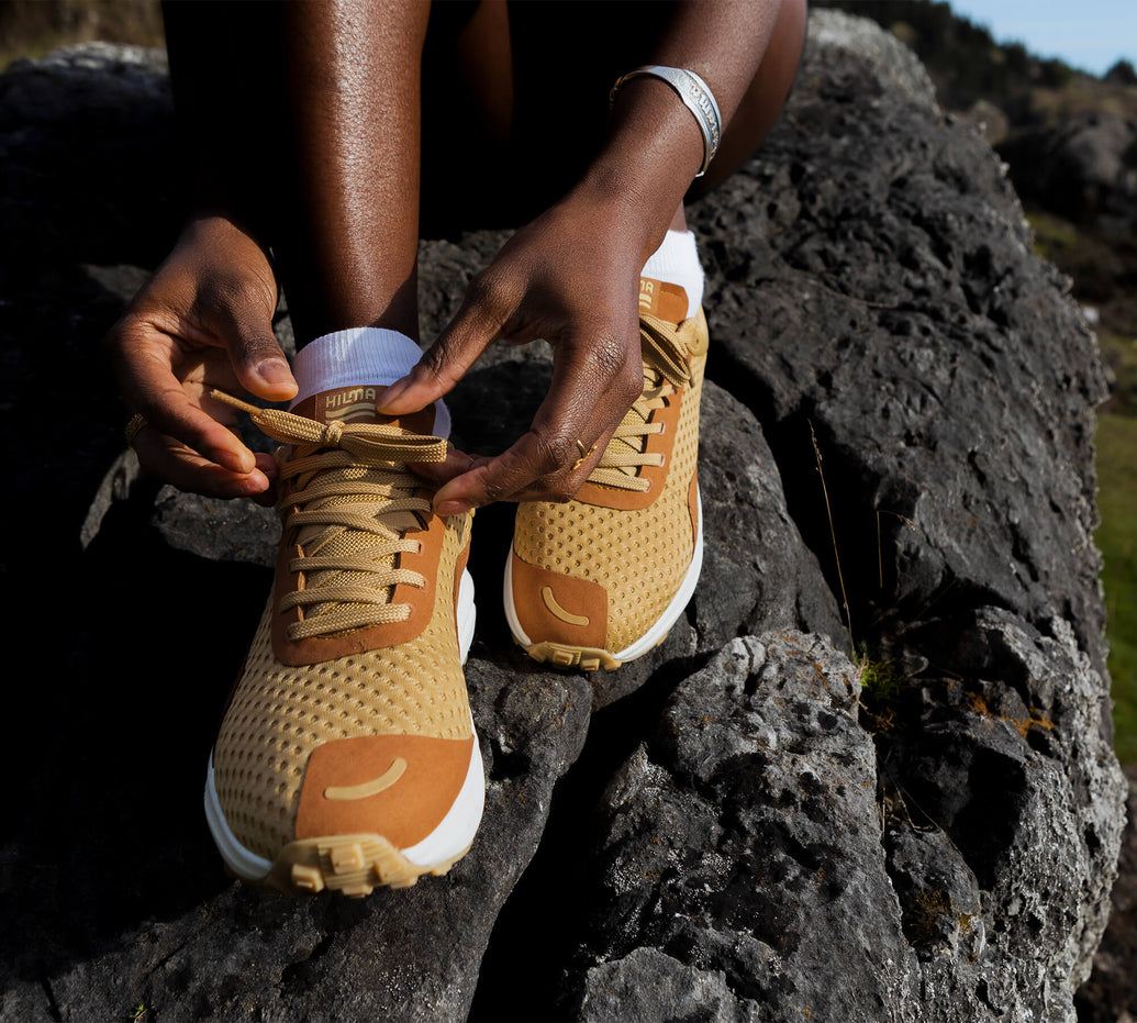 Person sitting on a large rock tying a pair of Hilma Running Shoes in Sandstone colorway 